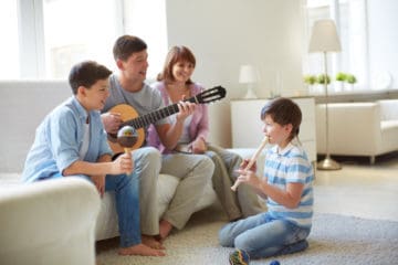 Portrait of siblings and their father playing musical instruments and their mom watching, happily. They are playing guitar, flute and a maracas, which is also called a musical shaker.