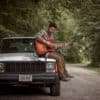 Man playing guitar on Jeep outside. Enjoying the outdoors in the woods.