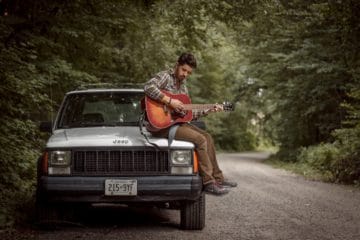 Man playing guitar on Jeep outside. Enjoying the outdoors in the woods.