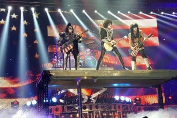 The band KISS on tall stage, playing guitar, with beam lights flashing Photo by John Pratt on Unsplash.