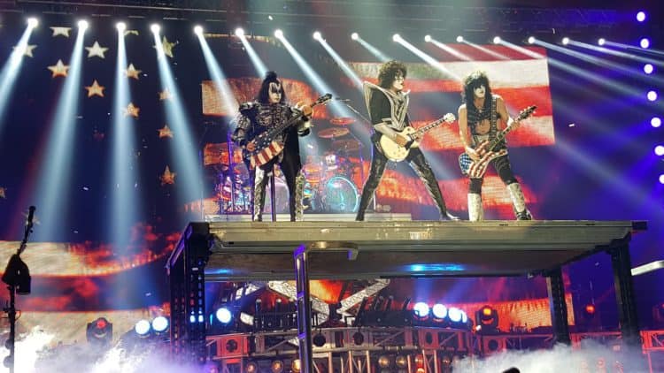 The band KISS on tall stage, playing guitar, with beam lights flashing Photo by John Pratt on Unsplash.