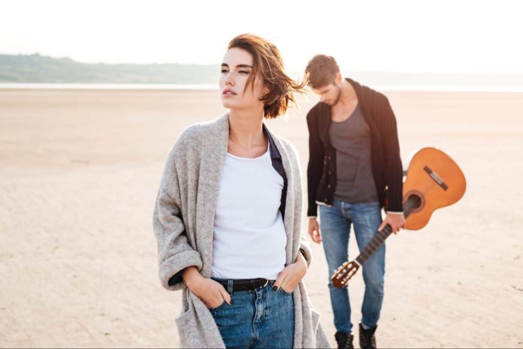 Man carrying guitar with head down. Woman in front, with hands in her pocket, looking side ways.