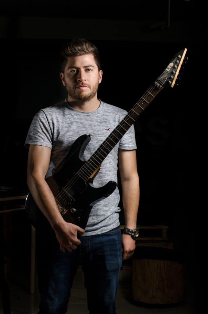 Man in grey shirt carrying guitar on the side of his body.