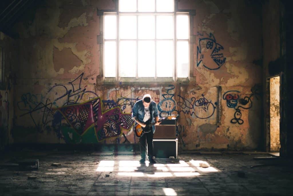 Guy playing guitar with an amp, in an abandoned, graffitied building, having fun.