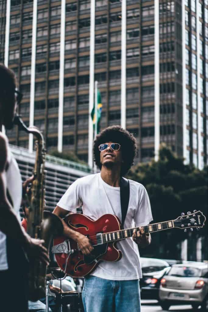 Man playing guitar with saxophone player outside, on a nice day. Also wearing sunglasses.