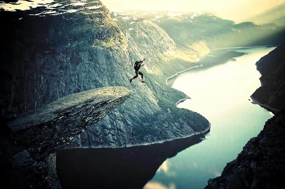 Man jumping off real high cliff, surrounded by mountains, to jump in the water. Has parachute.