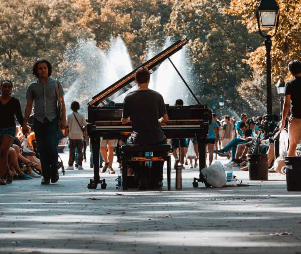 Man playing piano outside in the middle of the street, full of people.