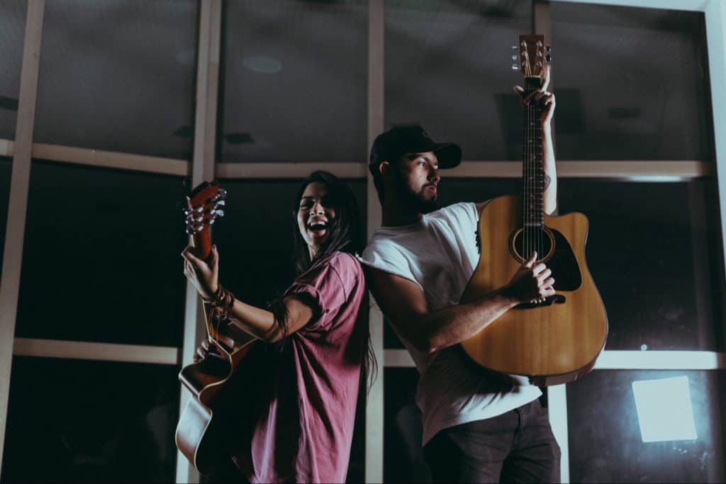 Man and woman playing guitar in front of windows.