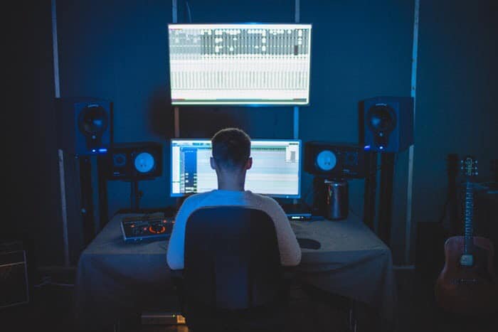 Young man sitting in studio room, looking at monitors and speakers. With guitar off to the side.