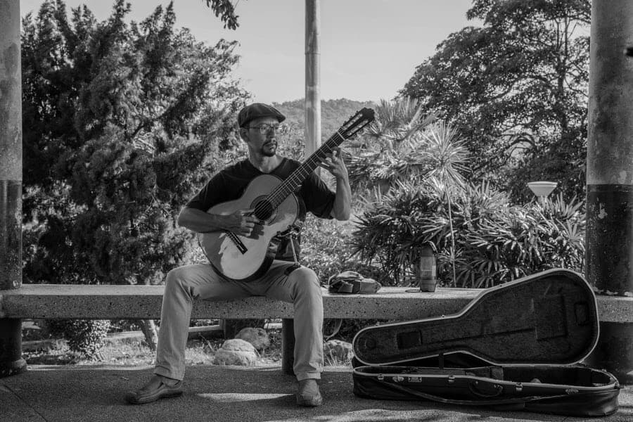 Playing the guitar outside, sitting down on a bench, on a beautiful day.