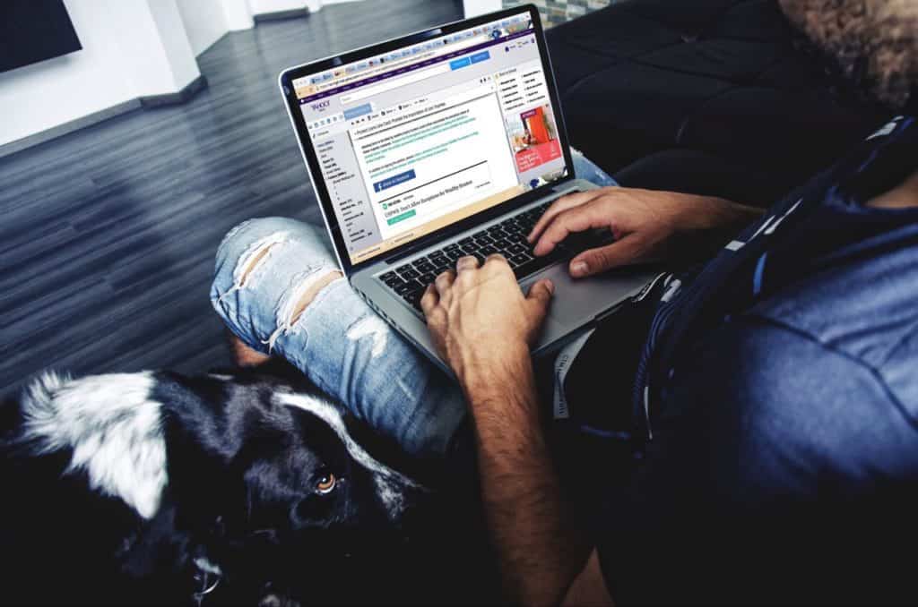 Person looking at emails, sitting down with laptop on his leg, with dog next to him.