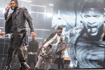 Usher dancing with hand to ear with musicians behind him and large projection screen behind him, of his projection image. Usher SAP Arena (cropped).jpg photo courtesy of D.roller.saparena. commons.m.wikimedia.org