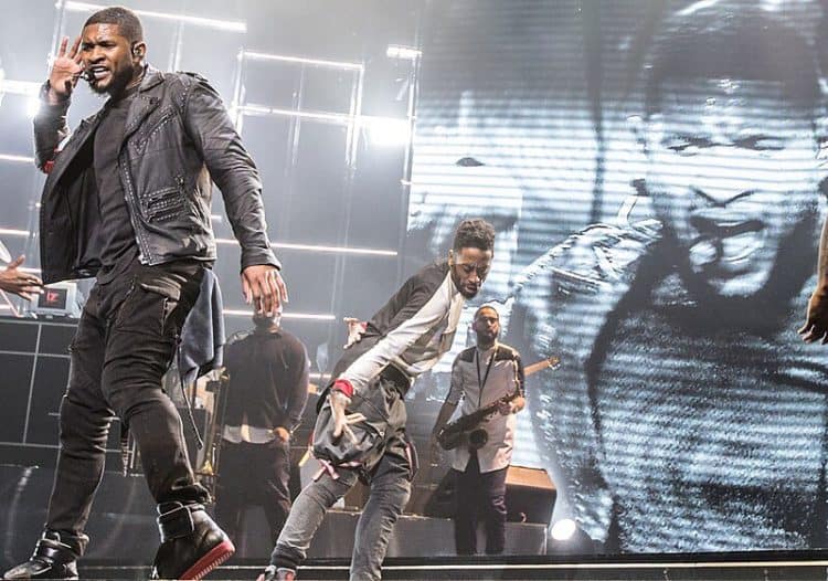 Usher dancing with hand to ear with musicians behind him and large projection screen behind him, of his projection image. Usher SAP Arena (cropped).jpg photo courtesy of D.roller.saparena. commons.m.wikimedia.org