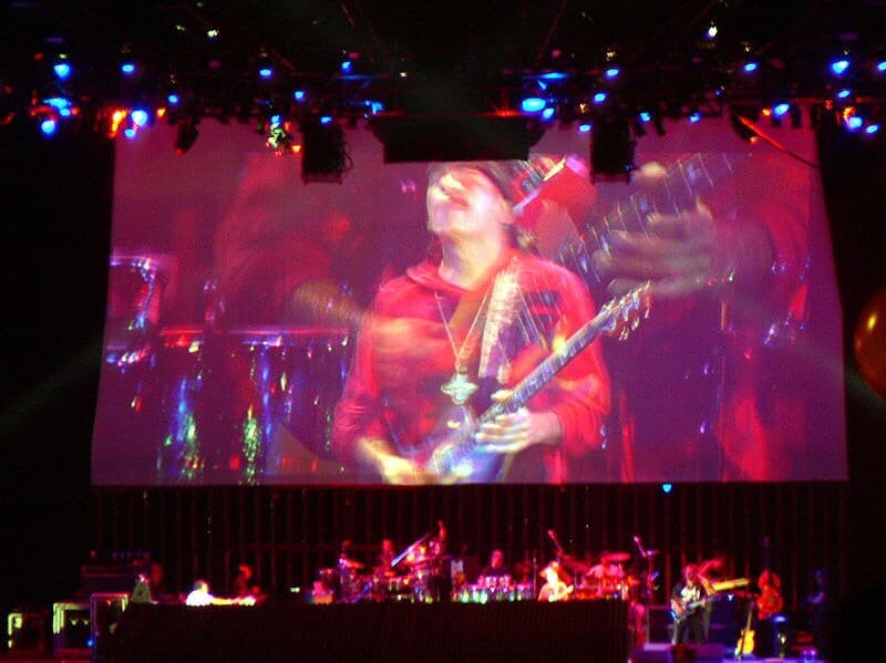 Carlos Santana on stage and on huge projection screen, filming him before a crowd.