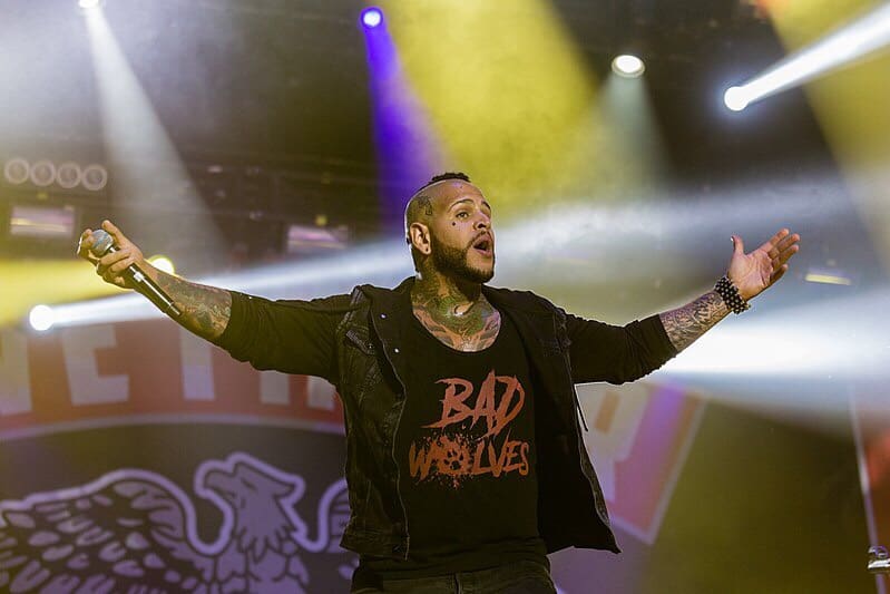 Tommy Vext holding up hands in the air with microphone in the other, with t-shirt that says, BAD WOLVES, Tommy Vext with Five Finger Death Punch photo courtesy of Alfred Nitsch. commons.m.wikimedia.org