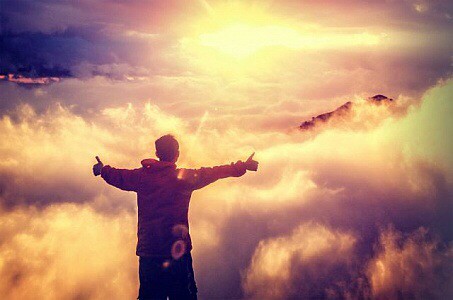 Kid with thumbs up in the heavens, clouds around him and mountains, Glorious bright light in front of him.