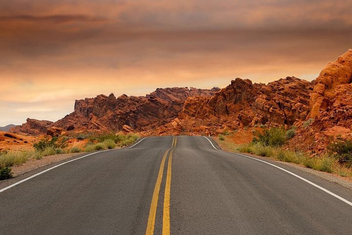 Street roadway outside, next to rocky mountains and green weed bushes, on a glorious looking, brownish, cloudy day.