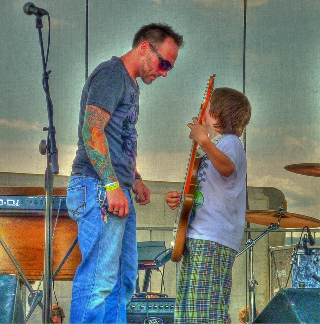 Man watching kid play guitar with careful, watchful eye, to make sure the kid is playing it right, having fun.