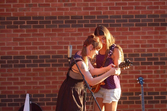 Girl showing another girl how to do chords on guitar by placing her hands on them, and the other girls hands on the guitar.