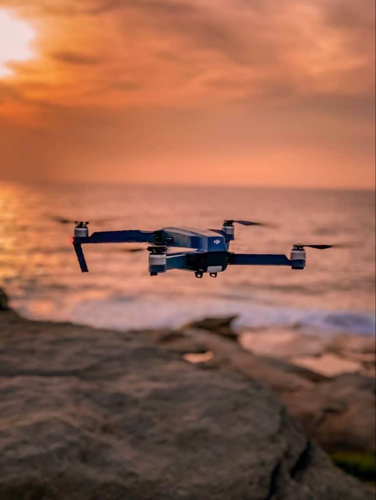 Blue drone flying by mountain boulders and ocean.