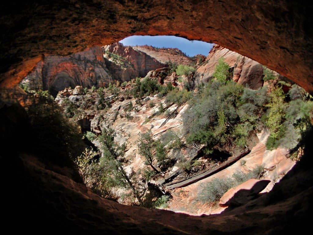 Bouldered cave with opening, showing green underbrush and rock mountains.