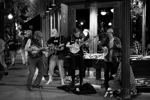 Men playing instruments outside busking. Two playing banjos, one playing guitar, the other playing a mandolin, at night.