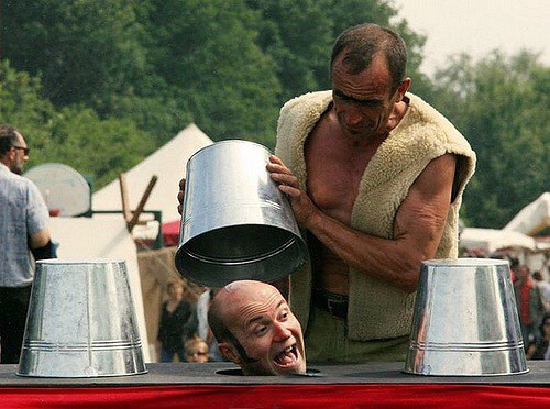 Clown, clowning around with buckets going on his head, by another man. And 2 other metal buckets around him. Clown has his head coming from out of the table, that has holes in it.