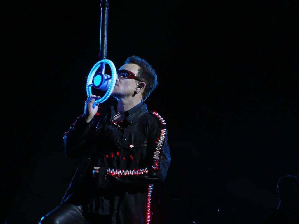 U2 singer, singing into lighted steering wheel microphone. Also has lighted up jacket on and sunglasses on.