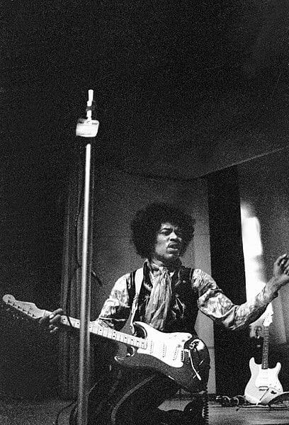 Jimi Hendrix on his knees, with guitar in one hand, with the other hand up in the air. Black and white picture.