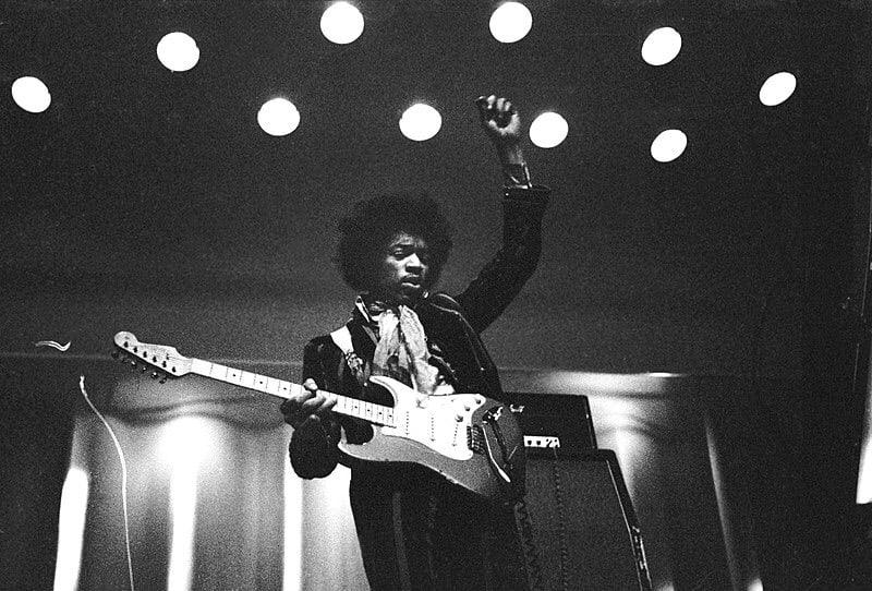 Jimi Hendrix strumming guitar and putting one hand up in the air, on stage. Black and white picture.