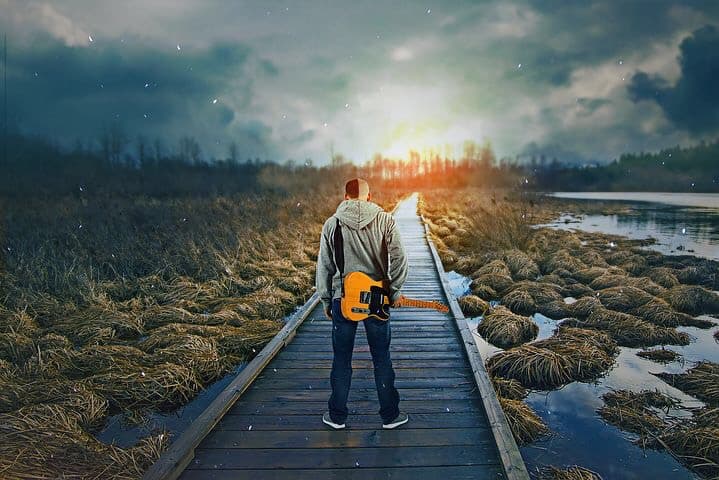 Man standing on a walking dock, on the water, with his guitar, with his head down.