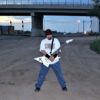 KBX under car overpass with trains in background, with guitar in his hands, striking a pose.