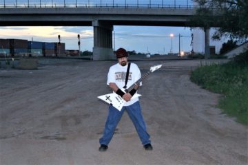 KBX under car overpass with trains in background, with guitar in his hands, striking a pose.