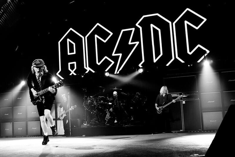 AC/DC musicians, on stage playing, black and white photo. Bright lights of AC/DC above them.